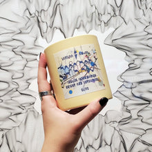 Load image into Gallery viewer, Hand holding a yellow candle with text in front of an art piece featured in MediaLive